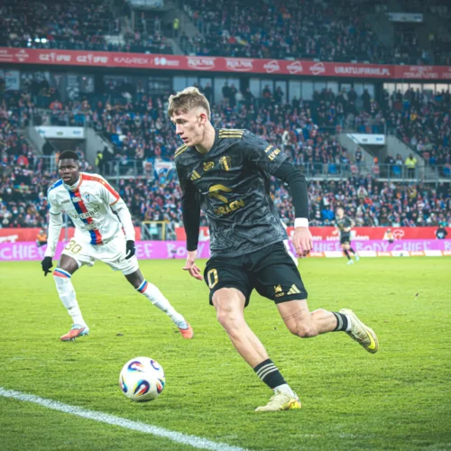 Na zdjęciu widać dwóch piłkarzy podczas emocjonującej akcji na boisku, z widownią w tle.