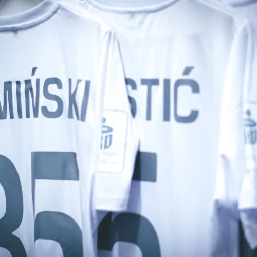 Na zdjęciu widoczne są trzy białe koszulki piłkarskie zawieszone na wieszaku, z widocznymi nazwiskami "KAMIŃSKI," "KOSTIĆ," i "VIĆ," oraz numerami na plecach.