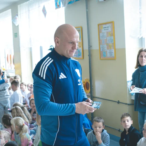 Na zdjęciu widoczny jest mężczyzna w niebieskim dresie, który trzyma telefon, oraz grupa dzieci siedzących na podłodze, a w tle znajduje się kobieta z dokumentami.