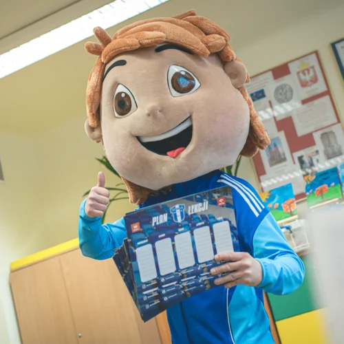 Na zdjęciu przedstawiony jest postać w dużej maskotce z uśmiechem, która trzyma planszę z planem lekcji i pokazuje kciuk w górę w kolorowej klasie.
