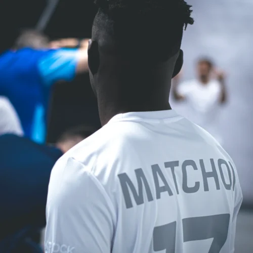 Na zdjęciu widoczny jest mężczyzna w białej koszulce z napisem "MATCHOI" i numerem 17, obserwujący inne osoby w tle.