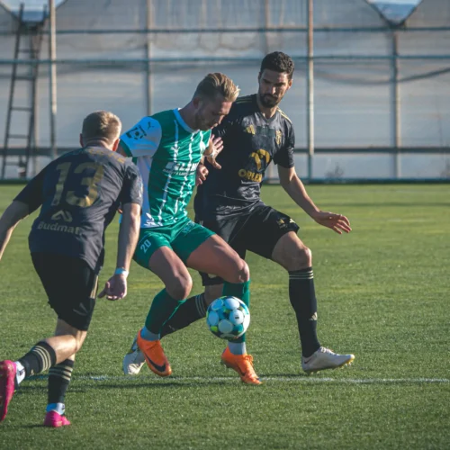 Na zdjęciu widać trzech piłkarzy rywalizujących o piłkę na boisku, z jednym zawodnikiem w zielonym stroju i dwoma w czarnych koszulkach.