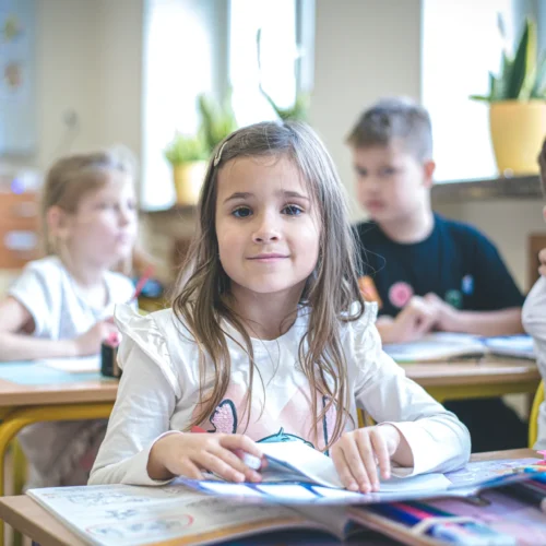 Na zdjęciu widać grupę dzieci w klasie, z uśmiechniętą dziewczynką z długimi włosami na pierwszym planie, która pracuje przy biurku.
