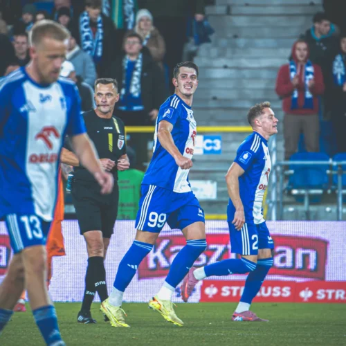 Na zdjęciu widać piłkarzy w niebiesko-białych strojach, którzy celebrują zdobycie bramki na tle kibiców na stadionie.