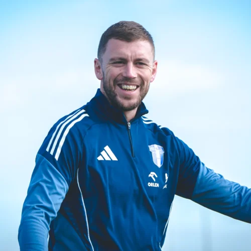 Mężczyzna uśmiecha się, nosząc sportową kurtkę w niebieskim kolorze z białymi detalami, na tle jasnego nieba.