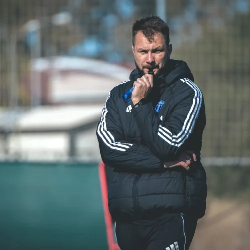 Trener w czarnej kurtce sportowej obserwuje trening piłkarski, trzymając dłoń przy ustach.