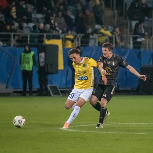 Na zdjęciu dwóch piłkarzy rywalizuje o piłkę podczas meczu na stadionie, z widownią w tle.