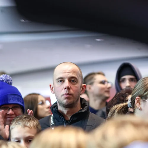 Na zdjęciu widać grupę ludzi, wśród których wyróżnia się mężczyzna z poważnym wyrazem twarzy, otoczony przez dzieci i innych uczestników zgromadzenia.