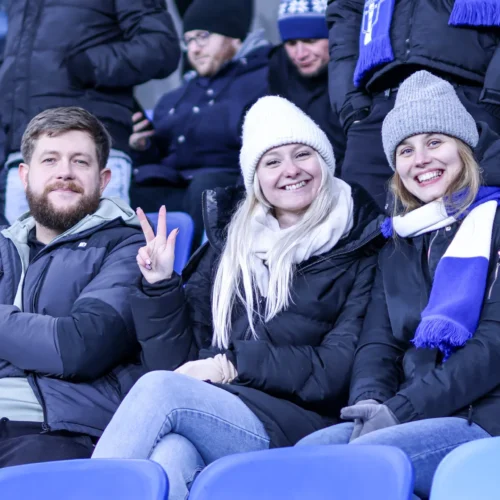 Na zdjęciu widać grupę trzech przyjaciół siedzących na trybunach, z uśmiechami na twarzach i w zimowych ubraniach, cieszących się atmosferą wydarzenia.