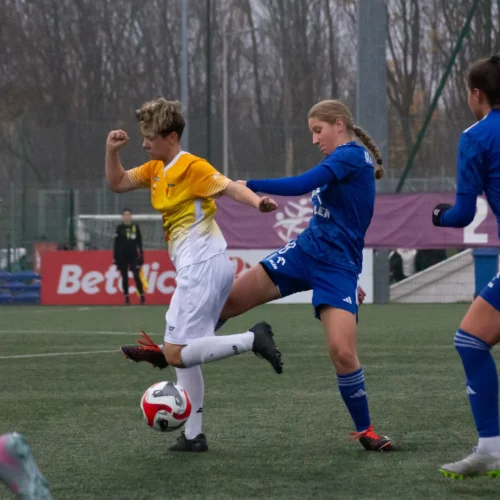 Na zdjęciu przedstawiona jest dynamiczna scena z meczu piłki nożnej, w której zawodniczka w żółtej koszulce stara się przejąć piłkę, podczas gdy przeciwniczka w niebieskim stroju podejmuje obronę.