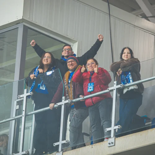 Grupa pięciu uśmiechniętych osób stoi na trybunie, ciesząc się z wydarzenia sportowego.