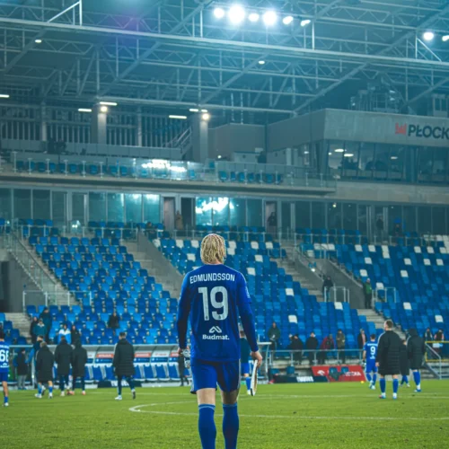 Na zdjęciu widoczny jest piłkarz w niebieskim stroju z numerem 19 na plecach, który idzie w kierunku zbrojowni na stadionie z pustymi trybunami w tle.