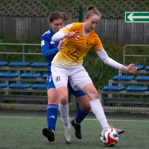 Na zdjęciu widać dwie zawodniczki grające w piłkę nożną, z jednej z nich wykonującej zwod przy próbie odbioru piłki przez przeciwniczkę.