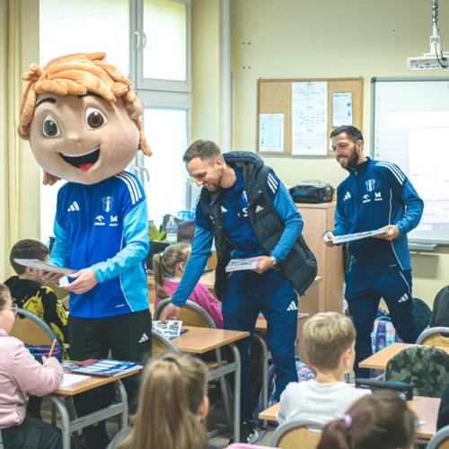 Na zdjęciu w klasie znajduje się grupa dzieci, z których jedno jest przebrane w kostium maskotki, a obok niego dwóch mężczyzn w sportowych strojach, przekazujących materiały uczniom.