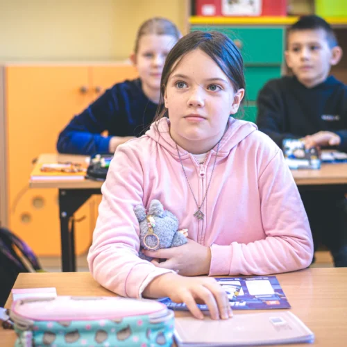 Na zdjęciu widać dziewczynkę w różowym swetrze, trzymającą pluszowego misia, siedzącą w klasie, a w tle widać innych uczniów.
