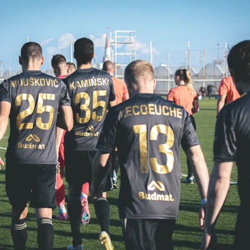Na zdjęciu widać grupę piłkarzy w czarnych strojach treningowych z widocznymi numerami, idących w stronę boiska.