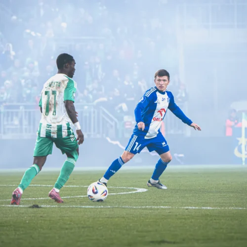 Mężczyźni w strojach piłkarskich grają na boisku otoczonym przez dym i z tłumem kibiców w tle.