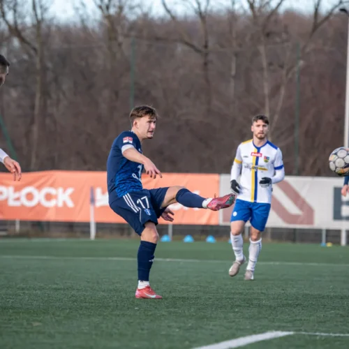 Na zdjęciu widać trzech piłkarzy na boisku, z których jeden wykonuje strzał, a dwaj inni obserwują akcję.
