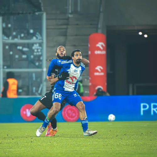 Na zdjęciu widać dwóch piłkarzy podczas dynamicznej akcji na boisku, gdzie jeden zawodnik trzyma drugiego, co sugeruje intensywną walkę o piłkę.