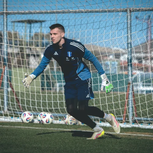 Na zdjęciu widać bramkarza w niebieskim stroju, który trenuje na boisku piłkarskim, w tle znajdują się piłki oraz bramka.