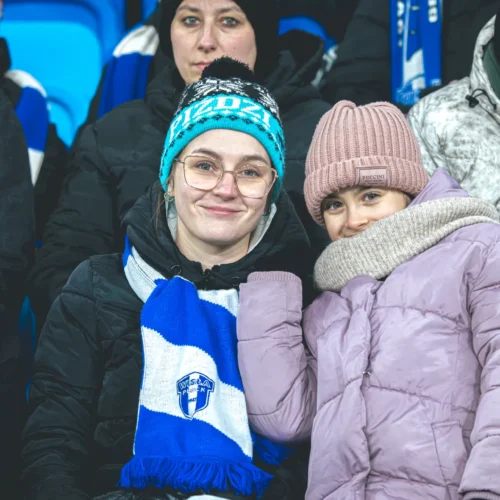 Na zdjęciu widać uśmiechniętą kobietę i dziewczynkę, obie w zimowych czapkach i kurtkach, siedzące na trybunach podczas meczu.