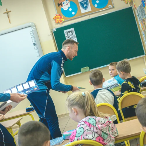 Dwóch mężczyzn rozmawia i rozdaje materiały dzieciom w klasie szkolnej.