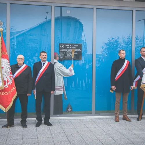 Grupa mężczyzn w formalnych strojach, trzymająca sztandary, stoi przed szklaną ścianą z tablicą upamiętniającą, podczas ceremonii.