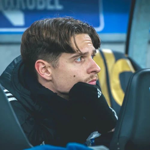 Mężczyzna w czarnej kurtce, z rękoma podtrzymującymi brodę, siedzi na ławce rezerwowych, koncentrując się na wydarzeniach na boisku.