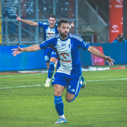 Piłkarz w niebieskiej koszulce cieszy się po zdobyciu gola, a w tle widać jubilującą postać kolegi z drużyny.