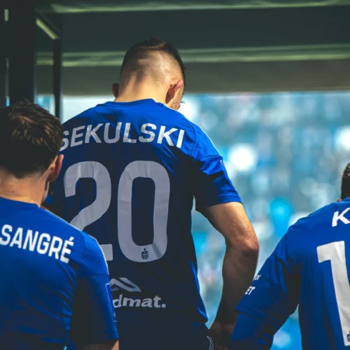 Fotografia przedstawia trzech piłkarzy w niebieskich strojach sportowych, przygotowujących się do wyjścia na boisko.