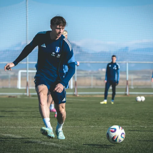 Zawodnik w niebieskim stroju piłkarskim kopie piłkę na boisku, podczas gdy w tle stoją inni gracze.