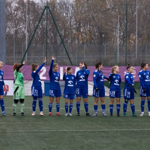 Na zdjęciu widoczna jest drużyna piłkarska kobiet w niebieskich strojach, stojąca w linii na boisku, podczas gdy zawodniczki machają w kierunku obserwatorów.