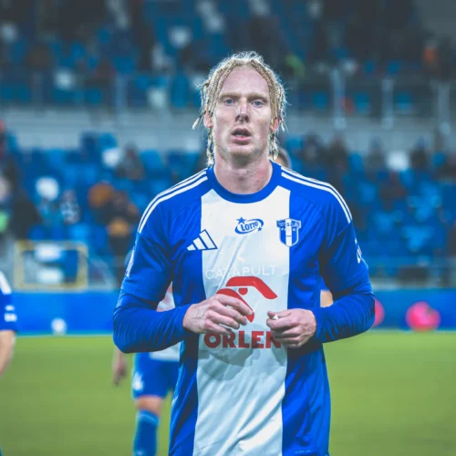 Na zdjęciu widać piłkarza w niebieskiej koszulce, który zmierza w stronę ławki rezerwowych na stadionie.