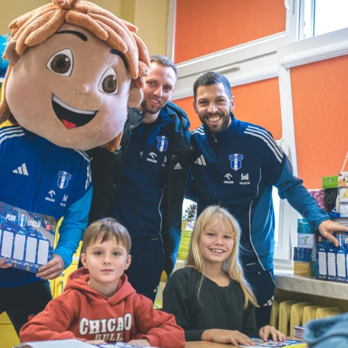 Na zdjęciu w klasie stoi postać z dużą głową oraz dwóch mężczyzn w uniformach sportowych, a przed nimi siedzą dzieci, które się uśmiechają.