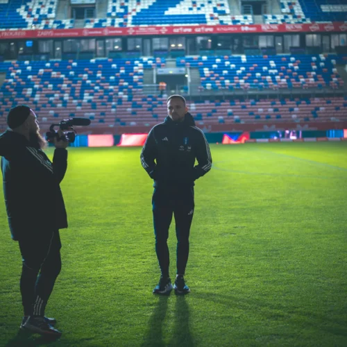Na zdjęciu widać dwóch mężczyzn rozmawiających na boisku, podczas gdy trzeci mężczyzna nagrywa ich wystąpienie kamerą.