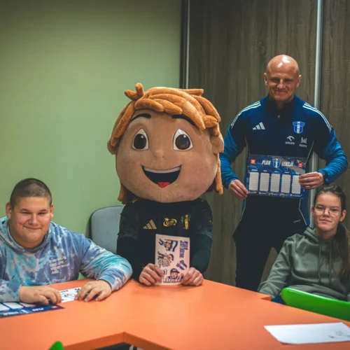 Na zdjęciu widoczna jest grupa osób, w tym jedna w kostiumie maskotki oraz dwie osoby siedzące przy stole, a także trener trzymający planszę z grafiką.