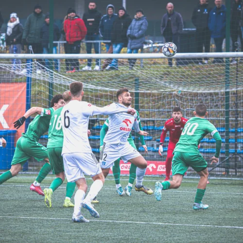 Na zdjęciu widać dynamiczną akcję w meczu piłkarskim, gdzie zawodnicy starają się złapać piłkę w pobliżu bramki, a na trybunach zgromadziła się widownia.