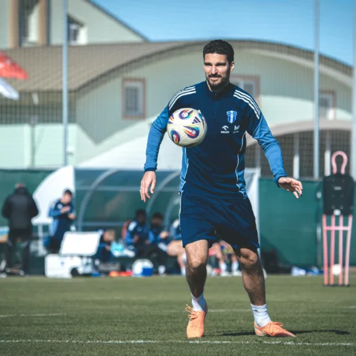 Na zdjęciu widać piłkarza ubranego w niebieski strój treningowy, który trenuje z piłką na boisku, z flagą w tle.