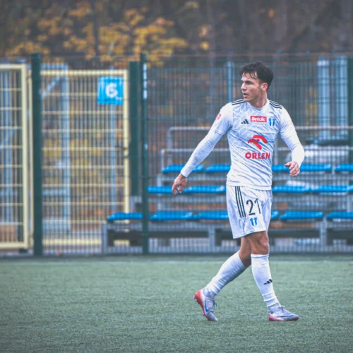 Na zdjęciu widoczny jest piłkarz w białym stroju sportowym, który spaceruje po boisku, na tle niebieskich trybun i ogrodzenia.