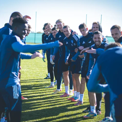 Kilkunastu mężczyzn w sportowych strojach stoi w rzędzie na boisku, z uśmiechami i gestykulującymi rękami, podczas treningu drużyny piłkarskiej.