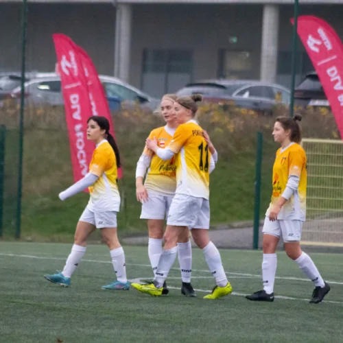 Na zdjęciu widać grupę kobiet grających w piłkę nożną, ubranych w pomarańczowo-białe stroje, które celebrują akcję na boisku.