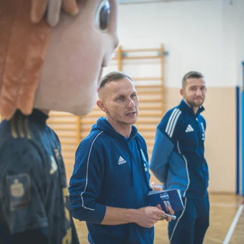 Na zdjęciu widoczna jest osoba w kostiumie maskotki oraz dwóch mężczyzn w dresach sportowych, z których jeden mówi, a drugi się uśmiecha, na tle sali gimnastycznej.