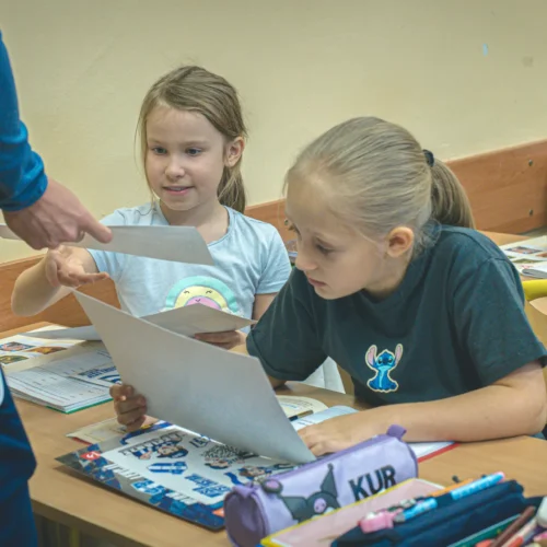 Na zdjęciu w klasie dzieci oglądają kartki papieru, a nauczyciel przekazuje im materiały do nauki.