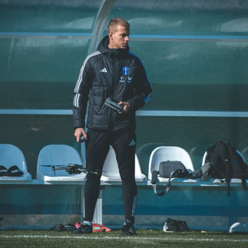 Na zdjęciu widać mężczyznę w kurtce sportowej, trzymającego drona, stojącego obok ławki na boisku, w otoczeniu opuszczonych rzeczy sportowych.