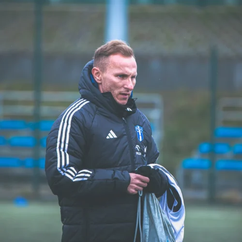 Na zdjęciu widać mężczyznę w czarnej kurtce treningowej z białymi detalami, trzymającego sprzęt na boisku sportowym.