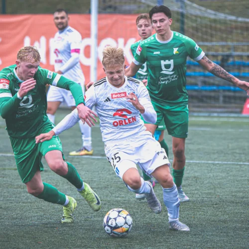 Akcja na boisku piłkarskim, gdzie zawodnik w białym stroju walczy o piłkę z przeciwnikiem w zielonym stroju, a w tle widoczni są inni gracze oraz baner reklamowy.