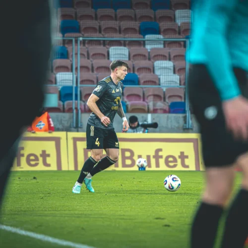 Na zdjęciu widoczny jest piłkarz przygotowujący się do rzutu wolnego na boisku, w tle widać widownię oraz sędziego.