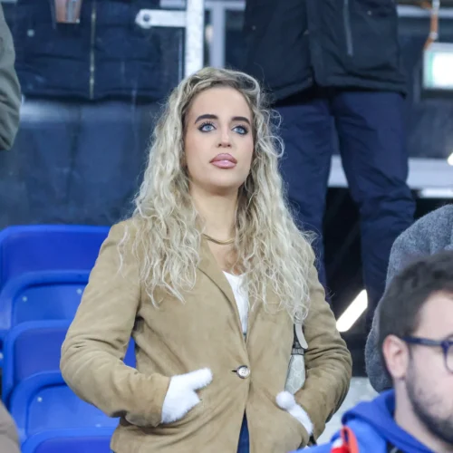 Kobieta z kręconymi blond włosami i niebieskimi oczami stoi wśród widowni, ubrana w beżowy płaszcz, z rękami schowanymi w kieszeniach.