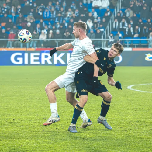 Na zdjęciu przedstawiono dwóch zawodników futbolowych w akcji, gdzie jeden z nich skacze, aby przejąć piłkę, a drugi próbuje go powstrzymać.