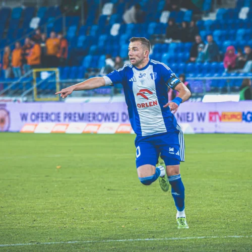 Na zdjęciu widać piłkarza w niebiesko-białym stroju, który cieszy się po strzeleniu gola, wskazując w stronę kibiców na trybunach.